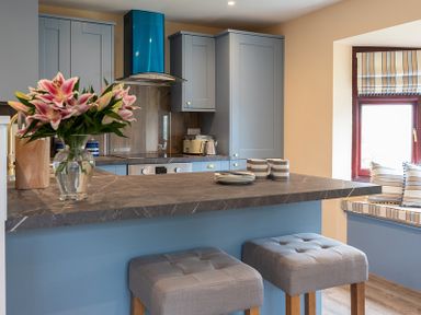 A kitchen with blue cabinets a marble countertop with flowers and plates two gray stools and a window seat with striped cushions at Brookside in Polzeath