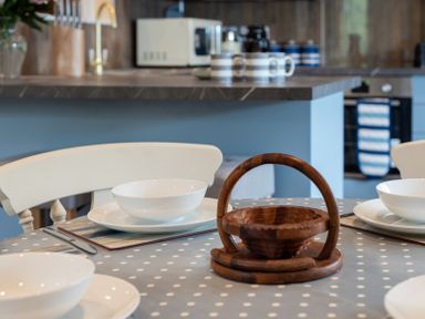 A kitchen dining table with white bowls and plates a wooden basket and chairs in a kitchen at Brookside Polzeath
