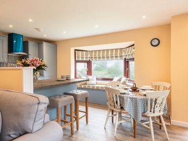 A kitchen and dining area with stools at the counter a round dining table with chairs and a bay window with seating at Brookside in Polzeath