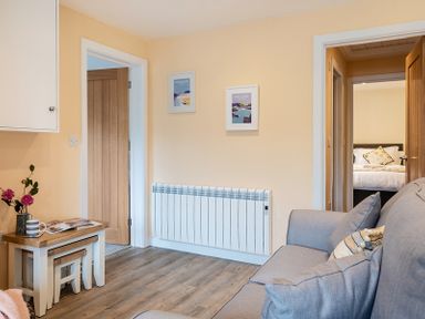 A room with a gray sofa and pillows a set of nesting tables with a cup and flowers on top two wooden doors and two framed pictures on a beige wall at Brookside in Polzeath