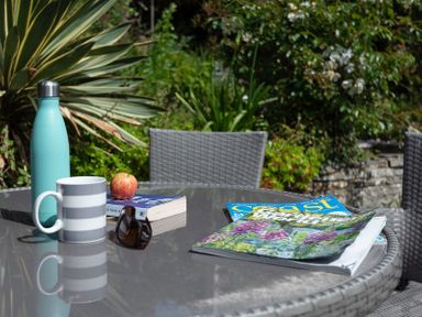 An outdoor glass table with a striped mug water bottle sunglasses book apple and magazines surrounded by wicker chairs and garden plants at Brookside in Polzeath