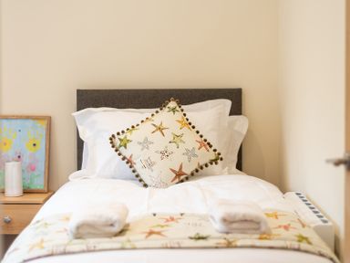A single bed with a starfish patterned pillow and blanket and white towels on it next to a wooden bedside table with artwork at Brookside in Polzeath