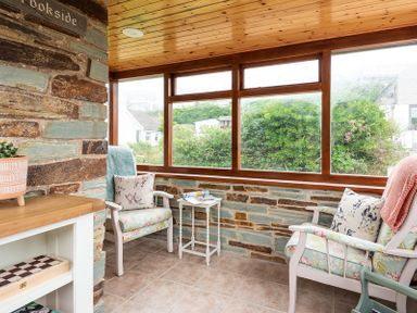 A sunroom with two armchairs a small white table and large windows showing greenery outside at Brookside in Polzeath