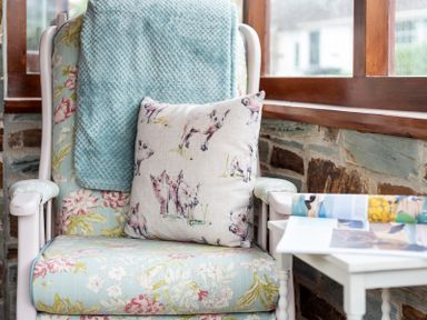 A floral patterned armchair with a blue blanket and a pig patterned cushion next to a stone wall and window at Brookside in Polzeath