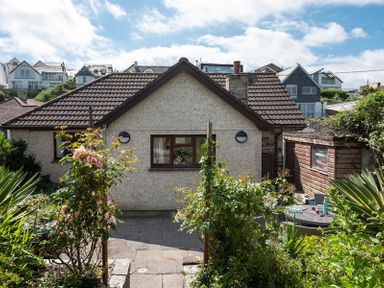 A garden with flowering plants and a paved patio in front of a single-story house with a tiled roof and a small shed at Brookside in Polzeath