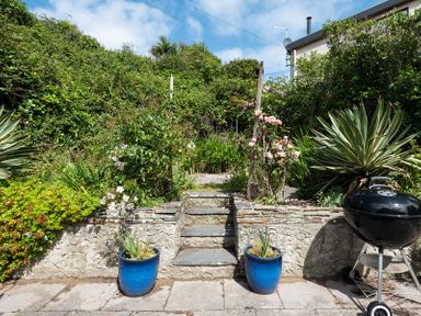 A garden with stone steps leading to a raised area flanked by blue plant pots and a black barbecue at Brookside in Polzeath