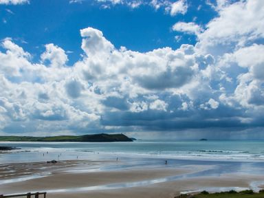 A beach with wet sand and a few people near the water with hills and an island in the background at Brookside in Polzeath