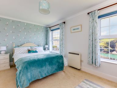 A bedroom with a bed covered in blue and white bedding two bedside tables with lamps two windows with curtains and a heater on the wall at Gulland in Port Isaac