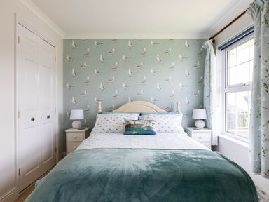 A bedroom with a bed, two bedside tables with lamps, a window with curtains, and seagull wallpaper at Gulland in Port Isaac