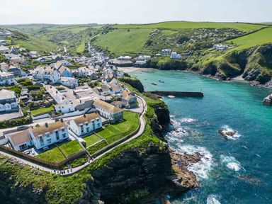 A coastal village with houses on cliffs beside a turquoise sea and green hills in the background at Gulland in Port Isaac
