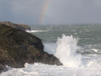 A coastal cliff with waves crashing against the rocks and a rainbow in the sky at Gulland in Port Isaac