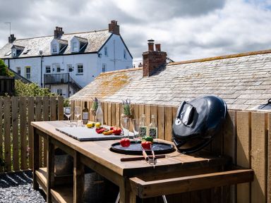 An outdoor wooden table with glass bottles, wine glasses, vegetables, and a black grill lid on a fenced patio at Mino Cottage in Port Isaac