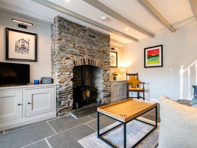 A living room with a stone fireplace a wooden coffee table a TV a chair with a cushion and framed artwork on the walls at Mino Cottage in Port Isaac