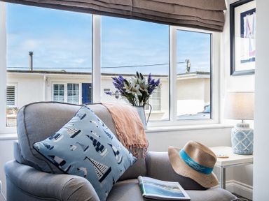 A gray armchair with a blue nautical pillow a hat a scarf and a book by a window with flowers and a table lamp at Mino Cottage in Port Isaac