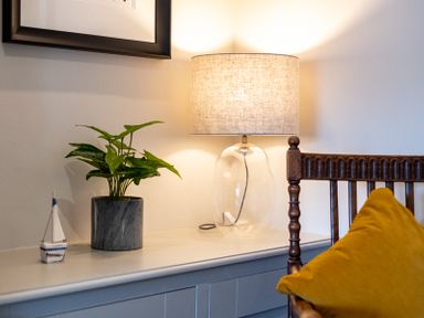 A tabletop with a potted plant a small sailboat model and a lamp next to a wooden chair with a yellow cushion at Mino Cottage in Port Isaac
