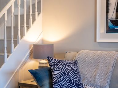 A sofa with decorative pillows and a throw blanket next to a lamp under a staircase at Mino Cottage in Port Isaac
