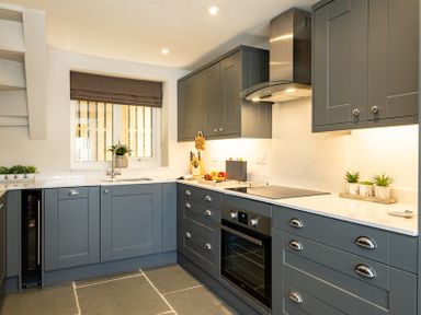 A kitchen with grey cabinets white countertops a stove and oven a window with a grey blind at Mino Cottage in Port Isaac