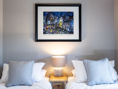 A bedroom with two beds separated by a wooden nightstand with a lamp and a framed painting above at Mino Cottage in Port Isaac