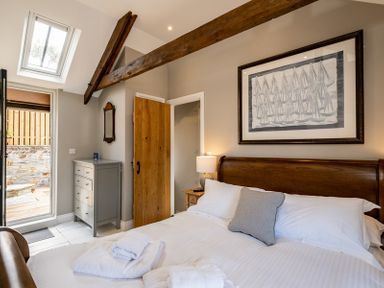A bedroom with a wooden bed a gray pillow white linens a framed artwork a wooden beam a gray dresser a wooden door and an open door leading outside at Mino Cottage in Port Isaac
