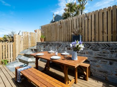 An outdoor dining area with wooden table and benches set with plates and glasses next to a stone wall and wooden fence at Mino Cottage in Port Isaac