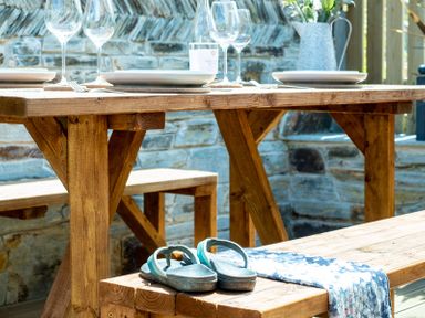 A wooden dining table set with plates and glasses and a bench with sandals and a blue cloth at Mino Cottage in Port Isaac