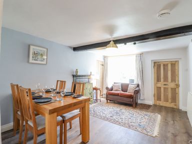 A dining area with a wooden table set with plates and glasses and a sitting area with a brown sofa and rug at Bluebird Cottage in Newmarket