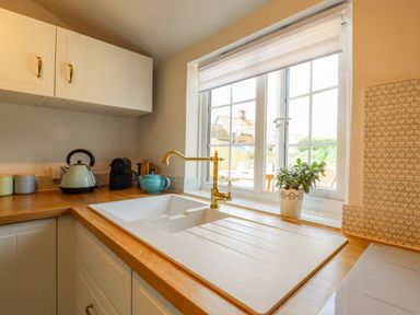 A kitchen sink with gold faucet under a window with a small plant and a kettle on the wooden countertop at Bluebird Cottage in Newmarket