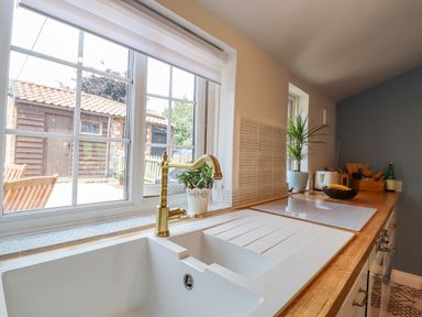 A kitchen countertop with a white sink and gold faucet a window showing an outdoor shed and a wooden chair and table at Bluebird Cottage in Newmarket