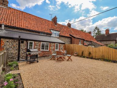 A backyard with gravel ground a wooden table and chairs and a barbecue grill near a stone house with red tiled roof Bluebird Cottage in Newmarket
