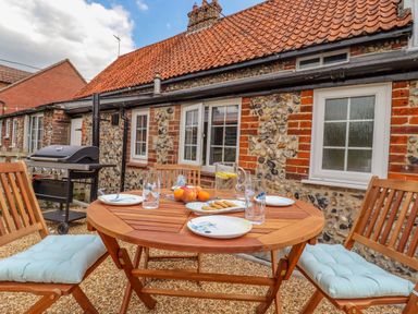 An outdoor patio with a wooden table and chairs set with plates glasses a pitcher of water with lemon and a barbecue grill at Bluebird Cottage in Newmarket