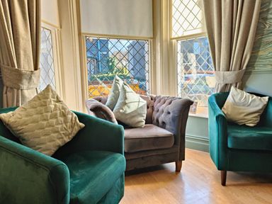 A sitting area with two green armchairs and a gray loveseat with cushions near leaded glass windows and beige curtains at Squirrel Lodge in Keswick