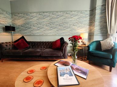 A living room with a sofa pillows a green armchair a table lamp flowers and a welcome book at Squirrel Lodge Keswick