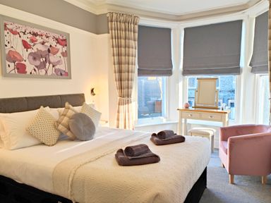 A bedroom with a bed with pillows and folded towels a dressing table and a pink armchair at Squirrel Lodge in Keswick