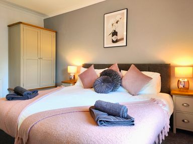 A bedroom with a bed featuring pink and gray cushions a pink blanket folded towels two bedside tables with lamps a wardrobe and a framed picture on the wall at Squirrel Lodge in Keswick