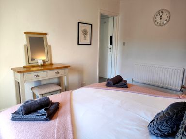 A bedroom with a bed covered in white and pink linens with folded towels on top a dressing table with a mirror a stool a wall clock and a framed picture at Squirrel Lodge in Keswick
