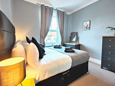 A bedroom with a bed, bedside lamp, window with curtains, dressing table, chest of drawers, and framed artwork at Squirrel Lodge in Keswick