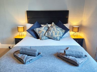 A bedroom with a bed with blue and patterned pillows two bedside tables with lamps and folded towels at Squirrel Lodge in Keswick