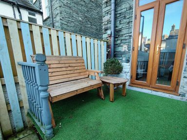 An outdoor seating area with a wooden bench and table a potted plant artificial grass and a wooden door at Squirrel Lodge in Keswick