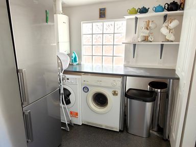A utility room with a refrigerator, washing machine, dryer, ironing board, iron, trash bins, and shelves with teapots and mugs at Squirrel Lodge in Keswick