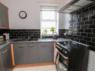 A kitchen with black tiled walls a window with plants on the sill a black stove and grey cabinets at Islestone 1 Temperance Terrace Berwick-Upon-Tweed