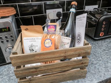 A wooden crate on a kitchen counter containing coffee packets biscuits cheese and a bottle near a toaster and microwave at Islestone 1 Temperance Terrace Berwick-Upon-Tweed