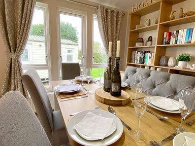 A dining area with a wooden table set with plates glasses and cutlery with chairs and a cushioned bench near windows and a bookshelf with books and decorative items at Meadow Lodge in Cayton