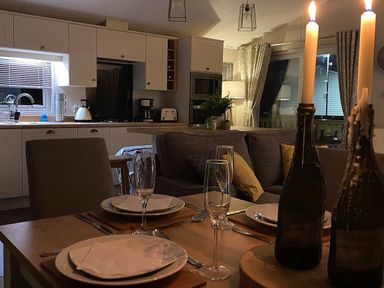 A dining table set with plates, napkins, glasses, cutlery, and two lit candles in bottles in a kitchen and living area at Meadow Lodge in Cayton
