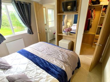 A bedroom with a bed a window with curtains a television a dressing table with flowers an open door showing a bathroom and another open door showing a closet at Meadow Lodge in Cayton