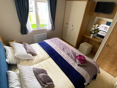 A bedroom with a double bed, pillows, folded towels, a wardrobe, a dressing table with a mirror and flowers, curtains on the window at Meadow Lodge in Cayton