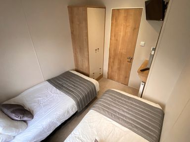 A bedroom with two single beds a wooden wardrobe a wooden door and a wall-mounted television at Meadow Lodge in Cayton