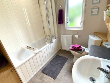 A bathroom with a bathtub and shower, a toilet with towels on the lid, a sink, a window, and shelving at Meadow Lodge in Cayton