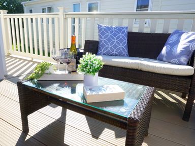 An outdoor seating area with a wicker sofa, cushions, a glass coffee table with a book, flowers, and wine glasses at Meadow Lodge in Cayton