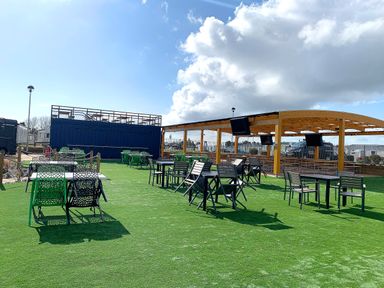 An outdoor seating area with tables and chairs on artificial grass with a covered wooden structure and large blue container in the background at Meadow Lodge in Cayton