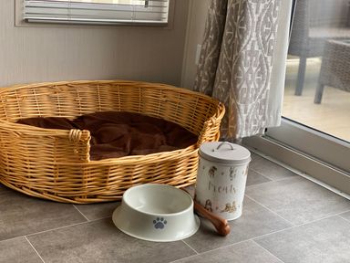 A pet bed with a cushion next to a food bowl and a treats container by a window with curtains at Meadow Lodge in Cayton